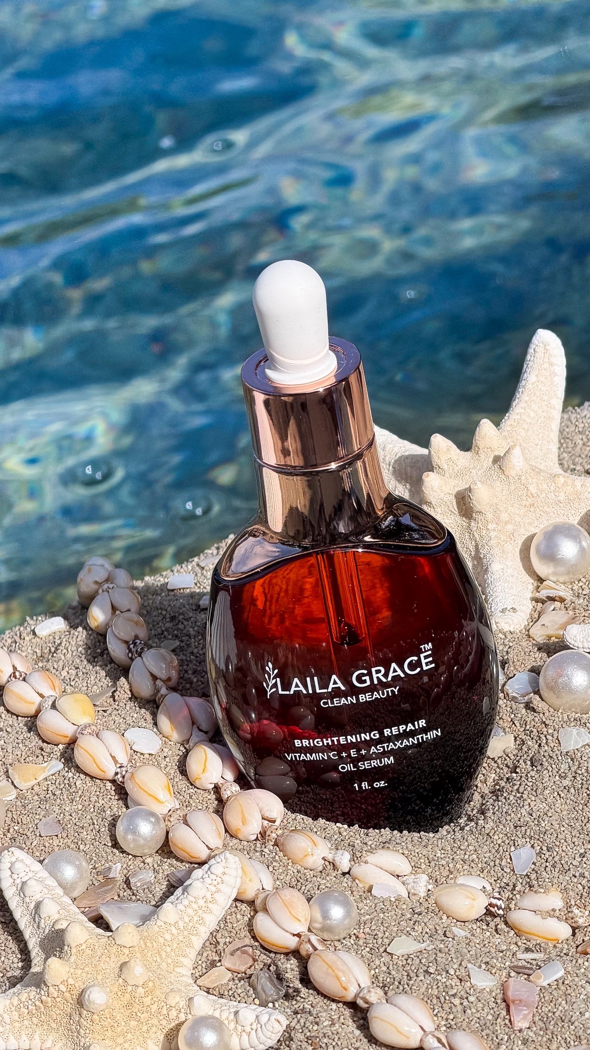 A bottle of Laila Grace Brightening Repair Vitamin C Serum sits on a sandy beach styled with starfish, shells, and pearls, with the blue ocean water in the background.