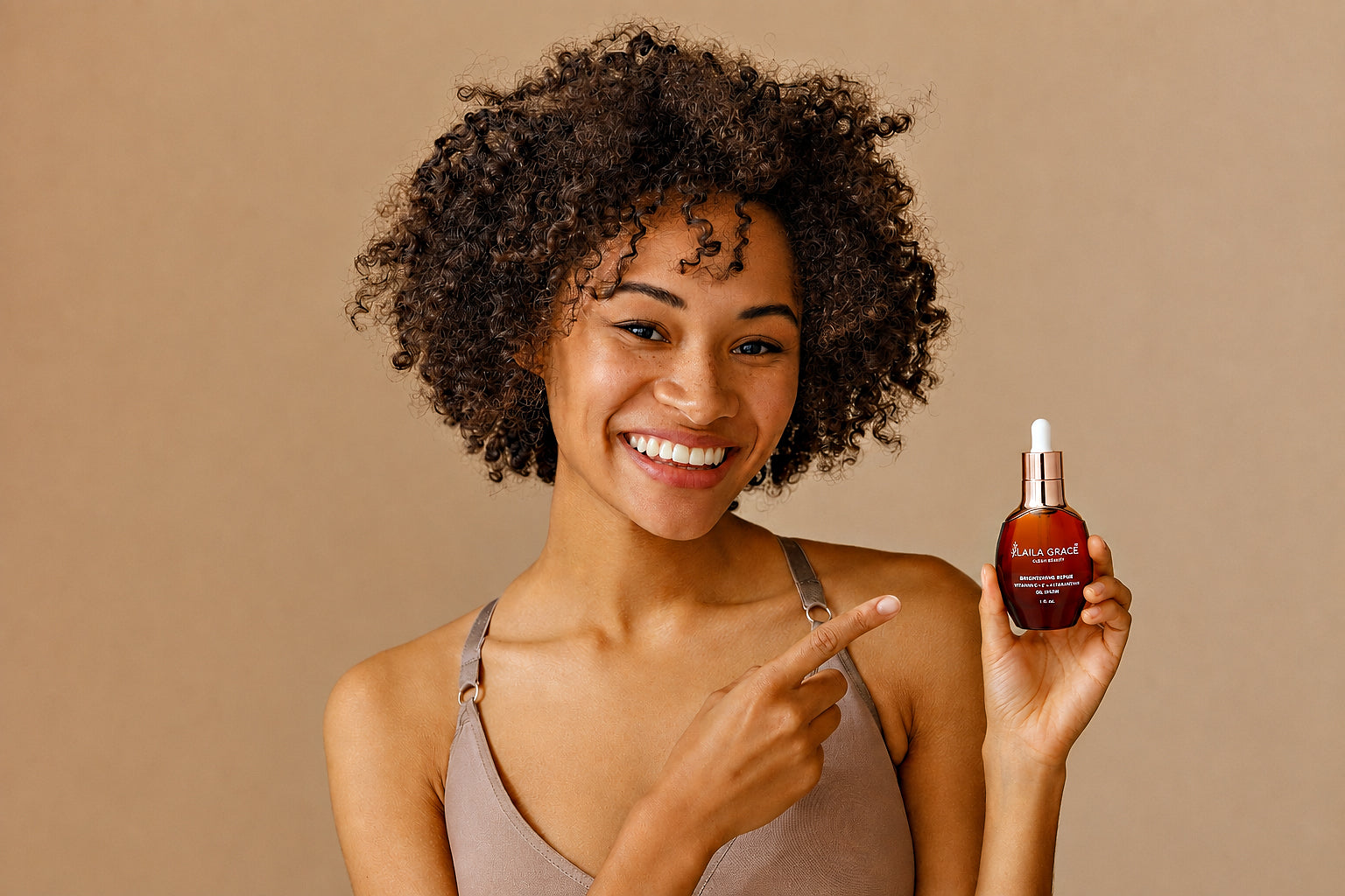 Woman with beautiful, glowing natural skin holding a bottle of Laila Grace Brightening Repair Serum and pointing to it against a beige background